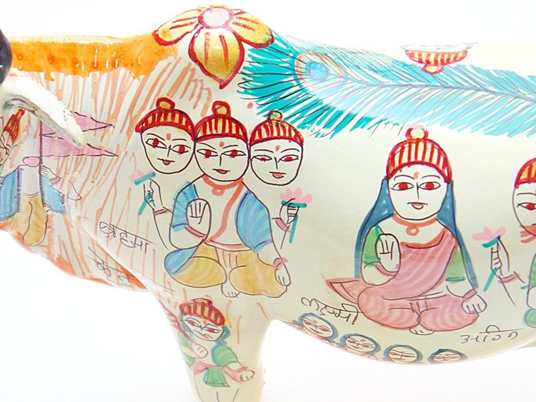 Close-up of cow idol body artwork, seated figures in red caps and blue-green clothing.